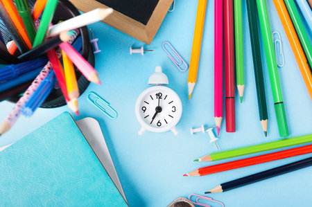 Back to school, Exam, Education concept on blue background. School scattered stationery, notepads, pens, pencils, paper clips and white alarm clock on blue table. Top view, copy space.の写真素材