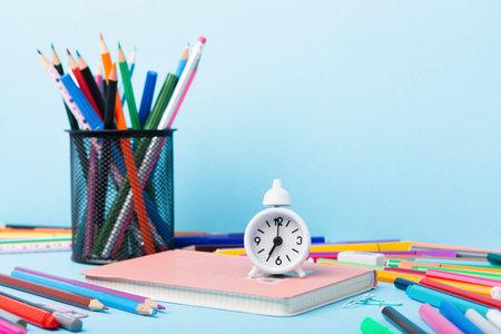 Back to school, Exam, Education concept on blue background. School scattered stationery, notepads, pens, pencils, paper clips and white alarm clock on blue table, copy space.の写真素材
