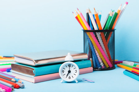 Back to school, Exam, Education concept on blue background. School scattered stationery, notepads, pens, pencils, paper clips and white alarm clock on blue table, copy space.の写真素材