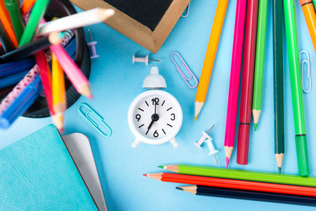 Back to school, Exam, Education concept on blue background. School scattered stationery, notepads, pens, pencils, paper clips and white alarm clock on blue table. Top view, copy space.の写真素材