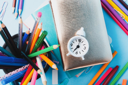 Back to school, Exam, Education concept on blue background. School scattered stationery, notepads, pens, pencils, paper clips and white alarm clock on blue table. Top view, copy space.の写真素材