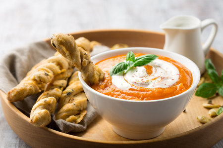 Homemade pumpkin soup with cream, fresh breadsticks and basil on rustic wooden background, selective focusの写真素材