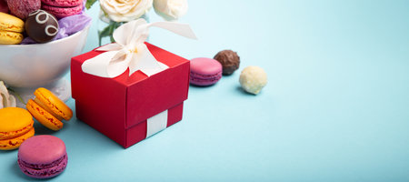 Sweet cookies macaroons, red gift box, rose flowers and chocolate candies on blue background. Presents concept for Mothers day, womans day, birthday, empty space for text messageの写真素材