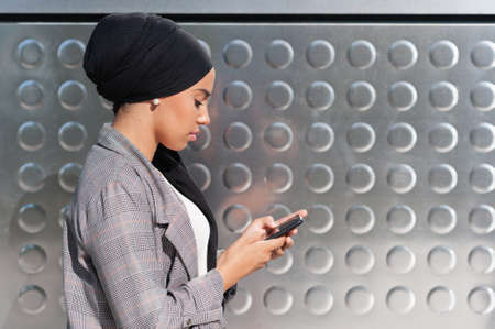 Side view of a Muslim woman standing using her phone with a wall of iron panels with circles patterns.の写真素材