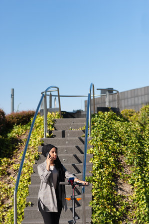 Concentrated Muslim woman with a black headscarf standing talking with her phone near a park stairs with an electric scooter in a sunny day.の写真素材