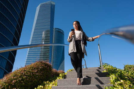 Vertical image of an adult woman wearing a black headscarf walking down on public staircase in the city using phone with buildings behind in a sunny day.の写真素材