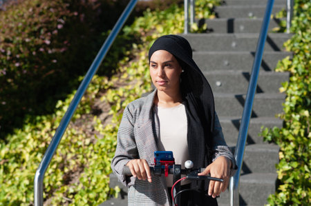 Front view of an adult muslim woman using her phone on an electric scooter in the park in a sunny day.の写真素材
