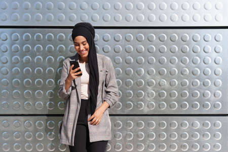 Front view of a cheerful Muslim woman standing looking and using her phone with a wall of iron panels with circles patterns behind.の写真素材