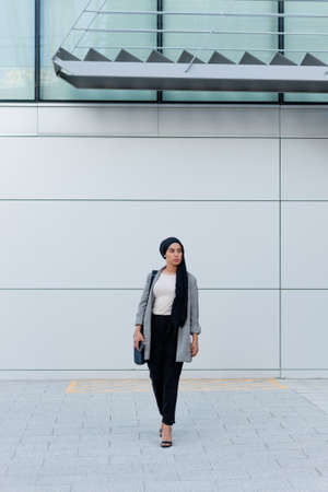 Full length view of an adult woman wearing a headscarf and holding her purse standing looking away against a building.の写真素材