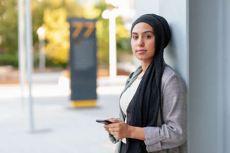 Front side view of a Muslim adult woman leaning on a beam column looking at camera while using her phone in a park.の写真素材