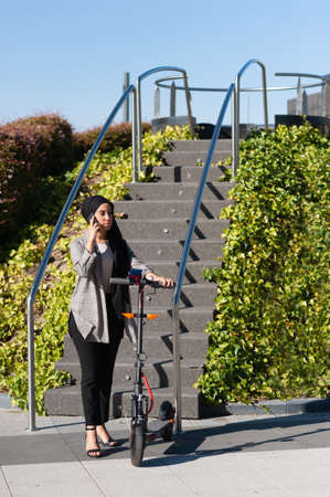 Concentrated Muslim woman with a black headscarf standing talking with her phone near a park stairs...