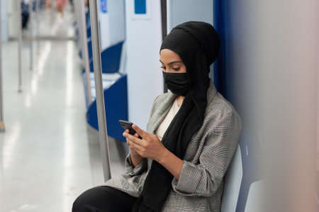 Muslim adult woman wearing a black face mask an a headscarf sitting on seat of subway while using her cellphone.の写真素材