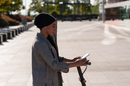 Side view of an adult muslim woman using her phone on an electric scooter in the park in a sunny...