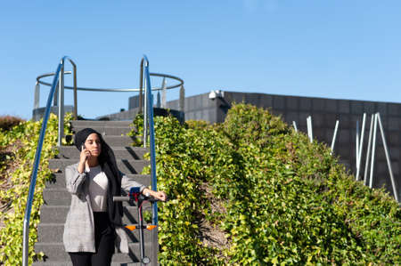 Concentrated Muslim woman with a black headscarf standing talking with her phone near a park stairs...