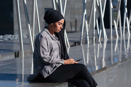 Side view of an adult woman wearing a black headscarf sitting on a modern marble structure using her...