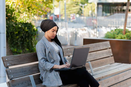 Front side view of an adult woman sitting on bench using her laptop in the park or city in a sunny day.の写真素材