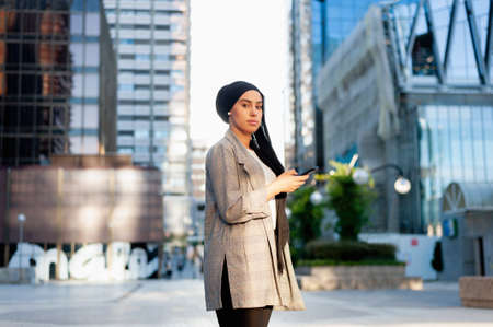 Side view of am unsmiling muslim woman standing using her phone in a urban area looking at camera in a sunny day.の写真素材