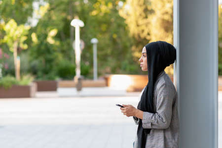Side view of a muslim adult woman leaning on a support beam column looking forward while using her phone in a park.の写真素材