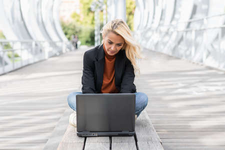 Frontal view of a blonde businesswoman sitting on a bench while working with a laptop in the streetの写真素材