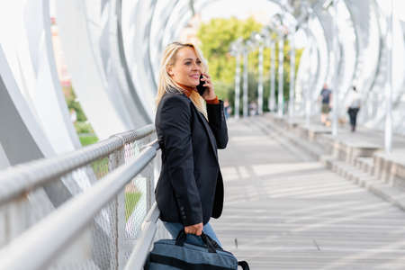 Smiling woman leaning on a railing of a urban bridge while talking with the mobile carrying a bagの写真素材