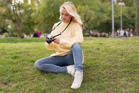 Blonde informal caucasian adult woman sitting on a park while using a camera hold by a leashの写真素材