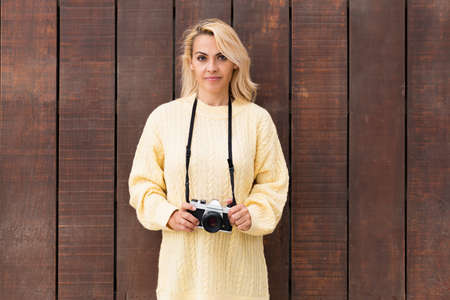 Portrait of a blonde woman holding a camera while facing the camera in front of a wooden wallの写真素材