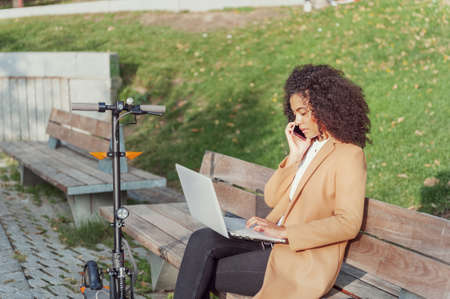 woman sitting on a park bench talks on the phone has a laptop and an electric scooter
