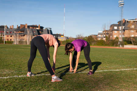 two young women doing stretching on the grass of a running track before playing sportsの写真素材