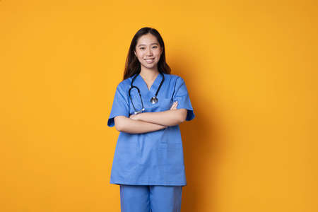 Studio photo with orange background of an asian doctor in uniform standing with her arms crossedの写真素材
