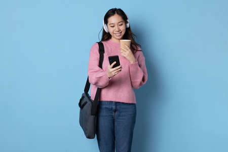 Studio photo with blue background of a woman using a mobile phone while holding a take away coffeeの写真素材