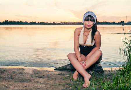 Vintage photo of relaxing young woman in nature. Retro fashion female sitting on lake shore and looking at cameraの写真素材