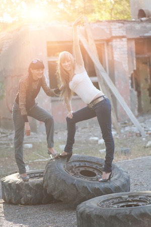 Brunette and blonde posing standing on giant tires outdoors at sunset backlit by sunの写真素材
