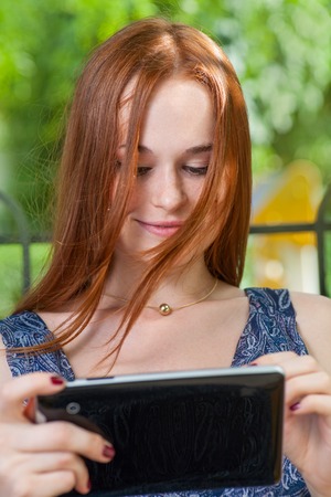 Redhead student leaning against a tree using her tablet on college campus. Redhead woman with tablet outdoorsの写真素材