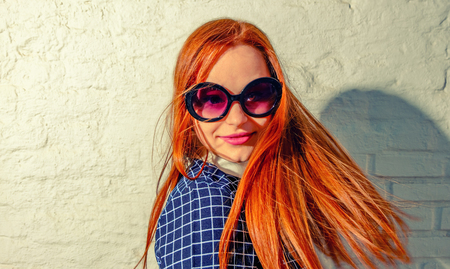 Trendy carrot-top girl turn around with her hair fly about her. Young woman weared in 60th fashion style sunglasses look back. A lot of copy space on white brick wall.の写真素材