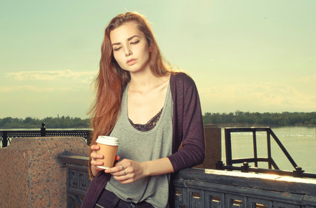 Portrait of beautiful brown haired girl standing and looking down. She keeping takeaway drink. Urban city scene. Outdoors portrait vintage film color imitation. toned image, colorizedの写真素材