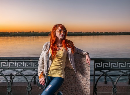 Redhair woman posing in sunglasses looking away of camera on river embankment, image with copy spaceの写真素材