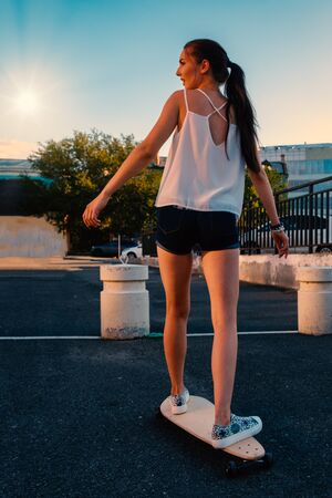 Beautiful girl riding skateboard in short denim shorts, view from back in front of early evening skyの写真素材