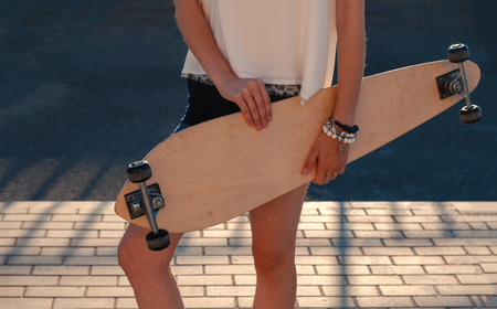 Front view of young girl in shorts holding longboard by both hands. Skateboard Extreme Sport Skater Activity Concept. Shadow pattern on pavement behind model.の写真素材