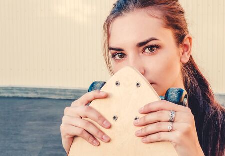 Girl holding skateboard in front of her face and looking at camera. A lot of space for text on wall behind the model.の写真素材