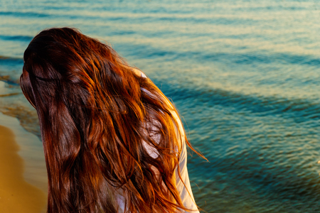 Girl with long red hair on the beach with her face hidden by hair, shot with copyspace on sea waterの写真素材