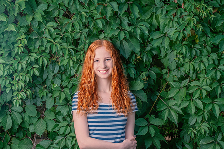 Cute young redhead woman standing in front of grape leaves wall looking up at the camera with a cheeky beaming smileの写真素材