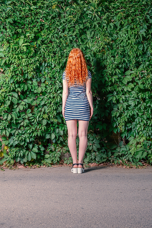 Rear view of Young redhead woman standing in front of grape leaves wall, full body shot