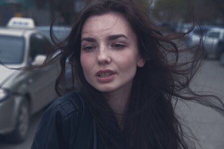 Sad girl with windy hair in the dark street in front of parked cars. Sadness and sorrow concept.
