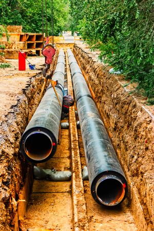 Two New heating water tube pipe in trench covered with black heat isulation, one of them hanging on cranes hookの写真素材