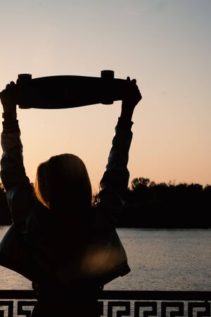 Woman posing with her hands holding skateboard raised up in the sunset sky, summer farewell concept. Vertical image.の写真素材