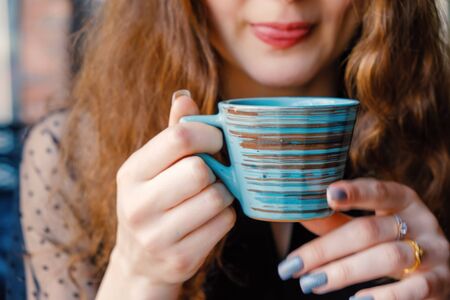 Curly girl with red hair poses in a cafe holding a Japanese-style cup at her juicy lips, she licks her lips while enjoying a drinkの写真素材
