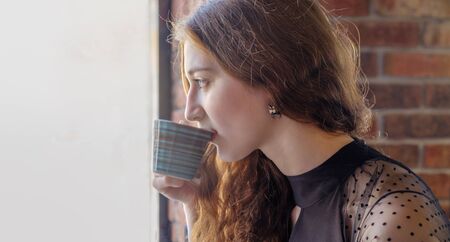 Curly girl with red hair poses in a cafe sipping from a cup of hot coffee, looking in profile with a...