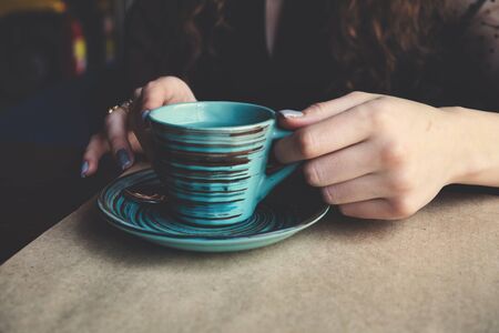 A close focus on the hands that hold a striped ceramic cup standing on a table covered with Kraft paper. Dark shadows fall on the hands and the cup, creating a slightly dark mood. The tinting imitates an old film photo. Copyspace is on the foreground.の写真素材