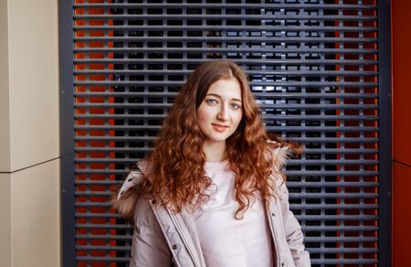Young redhead girl posing in front of the metal grid outside with her long curly hair spreaded all...