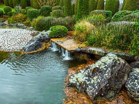 On the left side there is a mirror of water, and on the right there is a small waterfall framed by lush vegetation, and in the foreground there are large wild cobbles.の写真素材